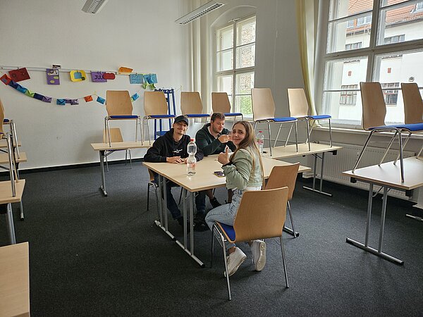 Podcast-Projekt: KAFFEE TRINKEN & SPIELEN Drei Schülerinnen und Schüler sitzen an einem Tisch und besprechen sich zu ihrem Projekt. Die Stühle der anderen Tische stehen den Tischen drauf.