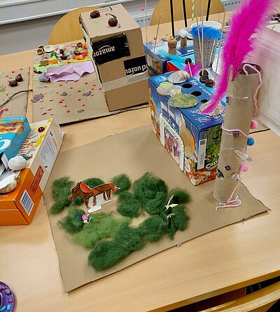 Auf einem großen Tisch stehen mehrere fantasievolle Landschaften aus Karton und Verpackungen; vorn ein brauner Pappboden mit grüner Wollwiese und ausgeschnittenem Pferd, daneben bunte Bastelobjekte und ein hoher Papprohr-Turm mit pinker Feder.