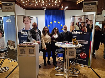 Zwei Schülerinnen und zwei Lehrer der Euro Akademie Bamberg präsentieren sich an ihrem Messestand zum Gruppenbild; auf den Tischen im Vordergrund liegen Infomaterial und ein Teddybär; im Hintergrund hängt eine Europaflagge.