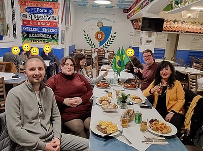 Mehrere Auszubildende der Euro Akademie Mainz sitzen gemeinsam an zwei rechteckigen Tischen in einem portugiesischen Restaurant in Mainz. Die Tische sind mit verschiedenen portugiesischen Gerichten gedeckt, darunter Pommes frites, gebratene Speisen und Salate. In der Mitte stehen zwei kleine grün-gelbe Brasilien-Flaggen. Die Wände sind blau-weiß gestrichen und mit Fußballschals portugiesischer Vereine sowie einem großen Wappen dekoriert, auf dem 'União Portuguesa Portugiese e.V.' steht. Im Hintergrund sind weitere Gäste, auf dem Bild unkenntlich gemacht, und Regale mit Weinflaschen zu sehen. Die Atmosphäre wirkt lebendig und gesellig.