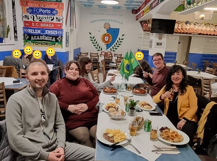 Mehrere Auszubildende der Euro Akademie Mainz sitzen gemeinsam an zwei rechteckigen Tischen in einem portugiesischen Restaurant in Mainz. Die Tische sind mit verschiedenen portugiesischen Gerichten gedeckt, darunter Pommes frites, gebratene Speisen und Salate. In der Mitte stehen zwei kleine grün-gelbe Brasilien-Flaggen. Die Wände sind blau-weiß gestrichen und mit Fußballschals portugiesischer Vereine sowie einem großen Wappen dekoriert, auf dem 'União Portuguesa Portugiese e.V.' steht. Im Hintergrund sind weitere Gäste, auf dem Bild unkenntlich gemacht, und Regale mit Weinflaschen zu sehen. Die Atmosphäre wirkt lebendig und gesellig.