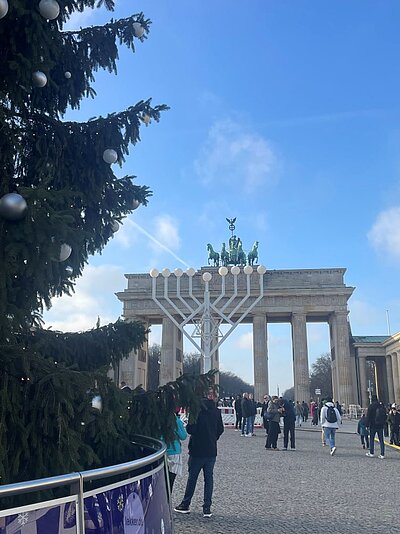 Szene im Freien mit einem großen, geschmückten Weihnachtsbaum im Vordergrund. Im Hintergrund erkennt man das Brandenburger Tor. Davor steht ein großes weißes Kunstwerk, welches den siebenarmigen Kerzenleuchter, die Menora, sowie den Davidstern als Symbole des Judentums darstellt. Menschen sind im Bereich des Bauwerks zu sehen. Der Himmel ist klar und blau.