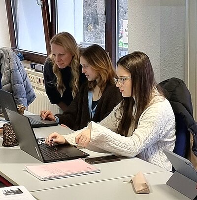 Zwei Personen sitzen zusammen an einem Tisch und arbeiten konzentriert an Laptops. Eine Person steht hinter ihnen und schaut auf die Bildschirme, möglicherweise um Unterstützung oder Anleitung zu geben. Der Raum ist hell und hat große Fenster, durch die Tageslicht einfällt. Auf dem Tisch liegen Notizblöcke, Stifte und persönliche Gegenstände wie ein Handy und eine Tasse.