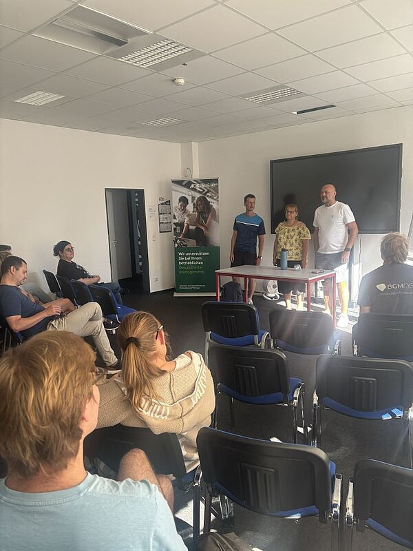 Drei Personen stehen vor einer Gruppe von sitzenden Teilnehmer*innen in einem Seminarraum. Die Atmosphäre wirkt wie eine Diskussion oder Präsentation. Im Hintergrund sind ein Flipchart und ein großer Bildschirm zu sehen.