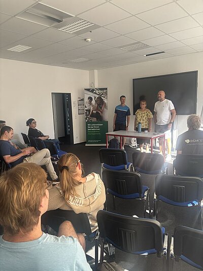 Drei Personen stehen vor einer Gruppe von sitzenden Teilnehmer*innen in einem Seminarraum. Die Atmosphäre wirkt wie eine Diskussion oder Präsentation. Im Hintergrund sind ein Flipchart und ein großer Bildschirm zu sehen.