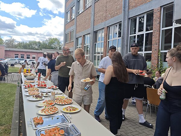 Ein langes Buffet mit Salaten, Obst und Snacks steht im Innenhof, während sich Teilnehmende bedienen und unterhalten.