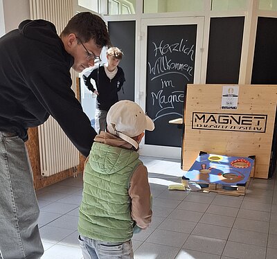 Ein Kind wirft bei einer Osteraktion in einem Eingangsbereich Säckchen auf ein bunt gestaltetes Wurfspiel, daneben unterstützt ein Jugendlicher.
