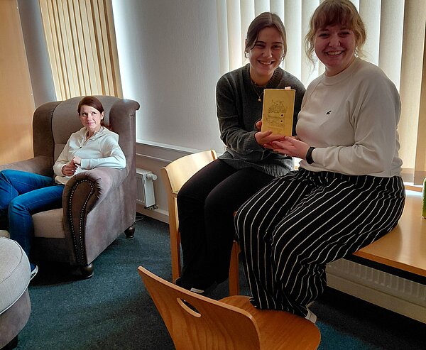 Zwei junge Frauen sitzen lächelnd auf einem Tisch und halten gemeinsam ein verziertes Windlicht in den Händen, im Hintergrund sitzt eine weitere Frau entspannt in einem Sessel.