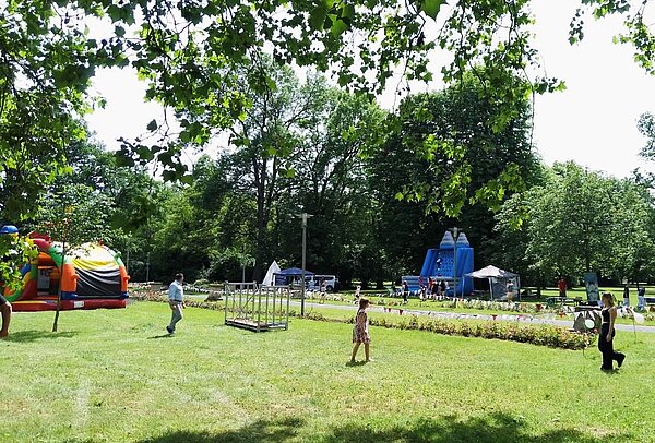 Ein grüner Park mit zwei aufblasbaren Spielgeräten (bunte Burg und blaue Rutsche) für Kinder. Im Vordergrund laufen zwei Kinder über den Rasen, im Hintergrund sind Bäume und weitere Besucher zu sehen.