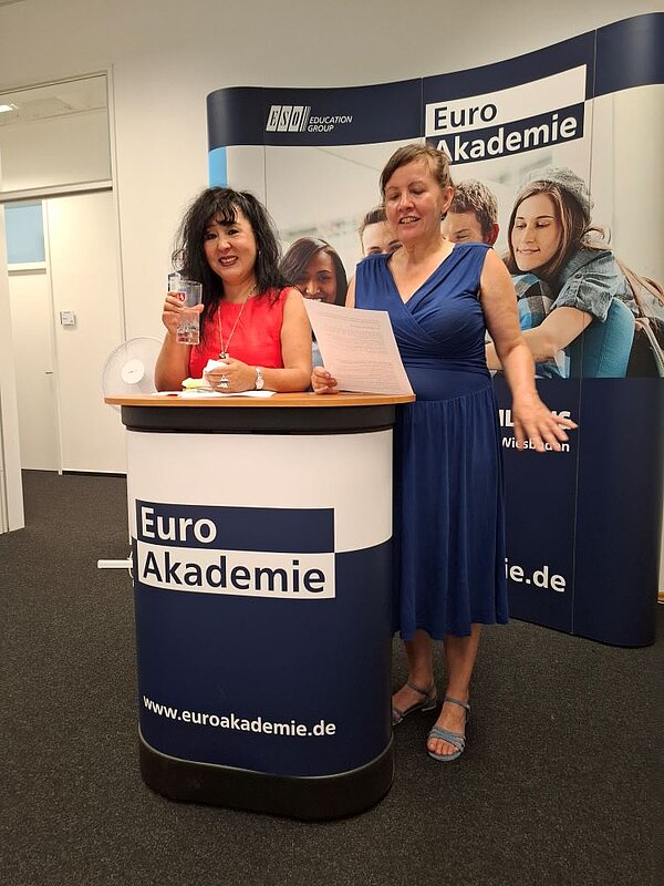 Zwei Personen stehen hinter einem Podium mit der Aufschrift "Euro Akademie". Eine Person hält ein Glas mit Wasser und lächelt, die andere Person scheint etwas vorzutragen. Sie hält ein Dokument in der einen Hand und gestikuliert mit der anderen. Sie trägt ein blaues Kleid.
