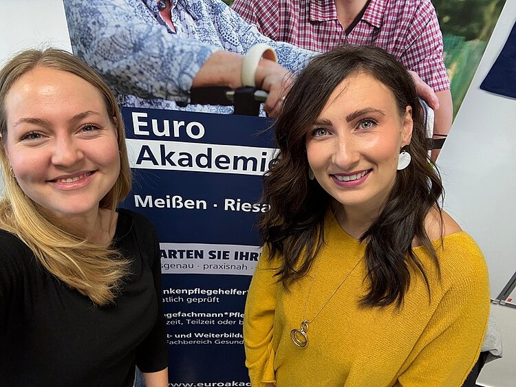 Zwei Mitarbeiterinnen der Euro Akademie Dresden stehen lächelnd vor einem Informationsbanner auf dem ‚Tag der beruflichen Schulen‘ im Berufsinformationszentrum (BiZ) am 15. November 2025. Das Banner bewirbt Ausbildungen und Weiterbildungen in den Bereichen Pflege und Soziales. Die Frauen wirken freundlich und aufgeschlossen und repräsentieren die Bildungsangebote der Euro Akademie. Die Szene vermittelt eine einladende und professionelle Atmosphäre.