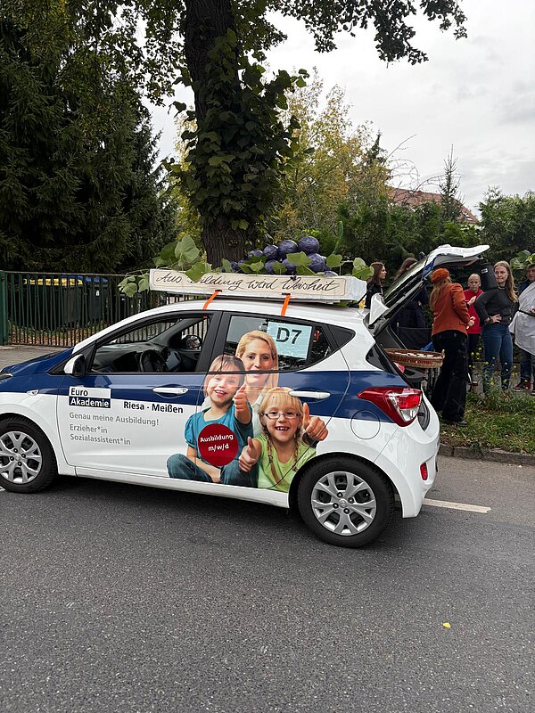 Ein weiß-blaues Auto mit Werbung der Euro Akademie Meißen steht mit offenem Kofferraum da. Auf dem Dach des Autos sind luftballongroße, selbstgebastelte Weintrauben und der Schriftzug "Aus Bildung wird Weisheit". Rechts hinter dem Auto stehen ein paar Menschen.