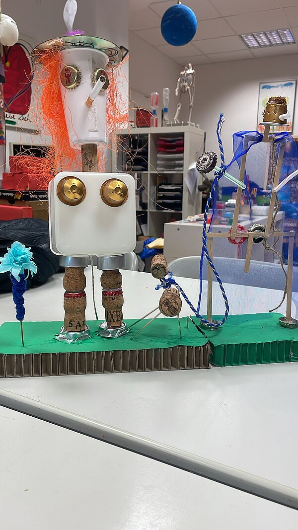 Eine Figur bestehend aus einem viereckigen Plastikbauch, einem Plastikbecher als Kopf, mit Korken als Füßen und Kronkorken als Augen und Brust, steht auf grüner Pappe.