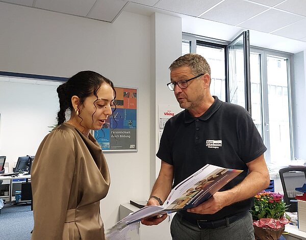 Ein Mann im schwarzen Polohemd zeigt einer Frau in brauner Bluse eine geöffnete Mappe, beide stehen im Büro.