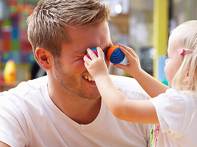 Ein Mädchen hält ihrem Kinderpfleger zwei bunte, kleine Bälle vor die Augen.