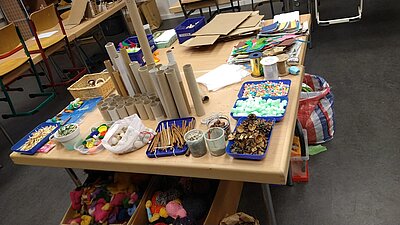 Ein großer Basteltisch ist mit vielfältigen Materialien gefüllt – darunter Papprollen, Federn, Kastanien, Stoffreste, Papier, Bänder und Naturmaterialien –, im Hintergrund sind Stühle und weitere Arbeitsplätze zu sehen.