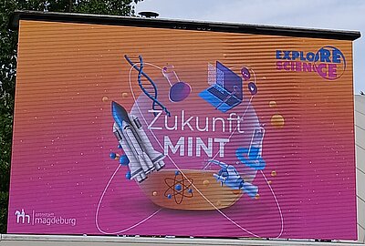 Ein Werbeplakat der Stadt Magdeburg mit dem Slogan „Zukunft MINT“ und dem Logo „Explore Science“. Das Plakat zeigt Illustrationen von Raketen, Atomen, Laptops und anderen wissenschaftlichen Symbolen.