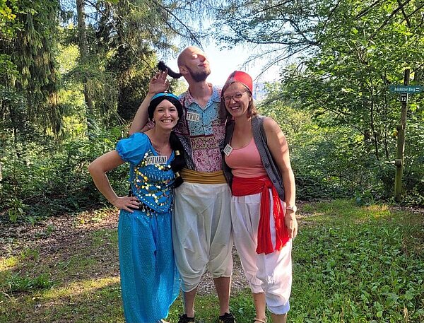 Zwei Frauen und ein Mann in orientalischer Verkleidung stehen in einer Waldumgebung und lächeln.