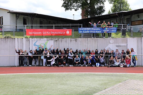 Auszubildende unterstützen Kreisjugendspiele Eine große Gruppe von Menschen steht für ein Foto vor einem Banner, auf dem “Kreisjugendspiele im Saale-Orla-Kreis” steht. Das Banner zeigt auch Logos von Sponsoren, darunter die Sparkasse. Die Gruppe befindet sich auf einem Sportgelände im Freien, und im Hintergrund sind Zuschauer zu sehen.