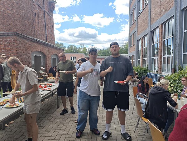 Zwei junge Männer stehen mit Tellern in der Hand vor dem Buffet eines Sommerfestes im Innenhof.