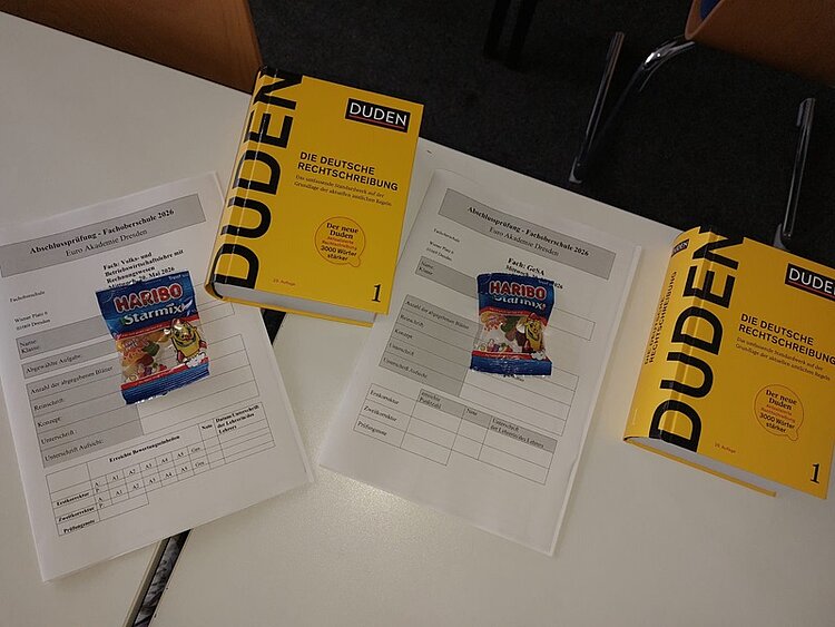 Auf einem Tisch liegen drei ausgefüllte Formulare, auf denen jeweils eine Tüte Fruchtgummis und ein gelbes Buch mit dem Titel 'Duden Die deutsche Rechtschreibung' platziert sind. Im Hintergrund sind Stuhlbeine und Teile von Stühlen zu sehen.