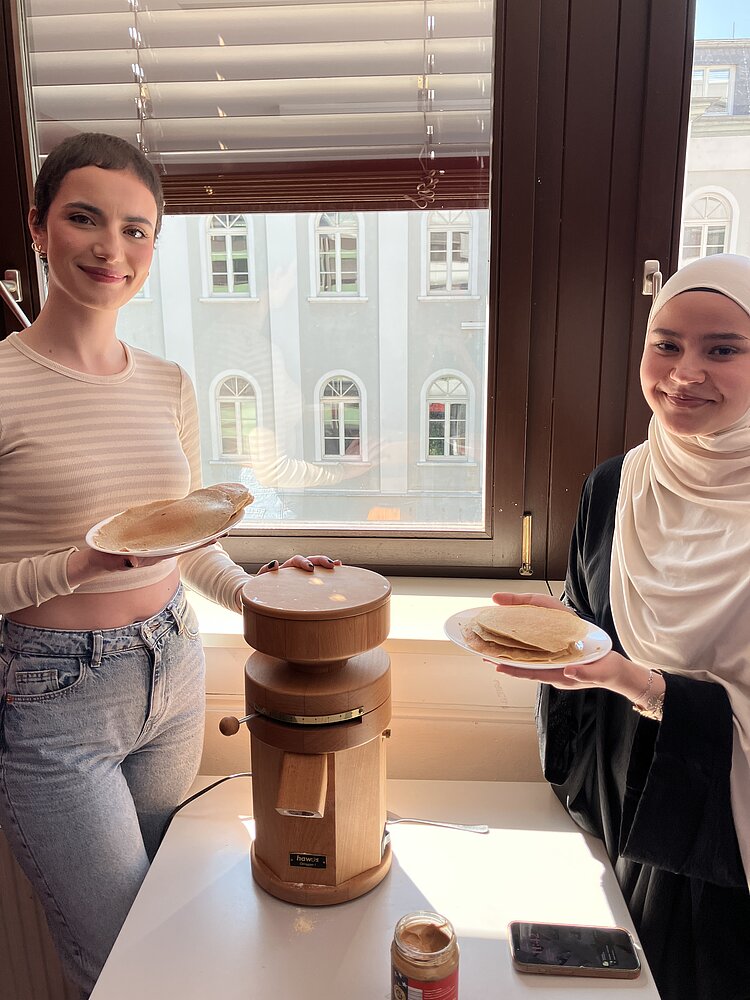 Zwei junge Frauen stehen in einem hellen Raum am Fenster. Vor ihnen eine Getreidemühle aus Holz. Jede hält einen Teller mit frisch gebackenen Pfannkuchen in der Hand und lächelt in die Kamera.