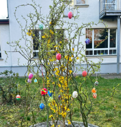 Alttext: Der gleiche Osterstrauch mit bunten Ostereiern, fotografiert aus einem leicht anderen Blickwinkel, vor dem gleichen Gebäude.
