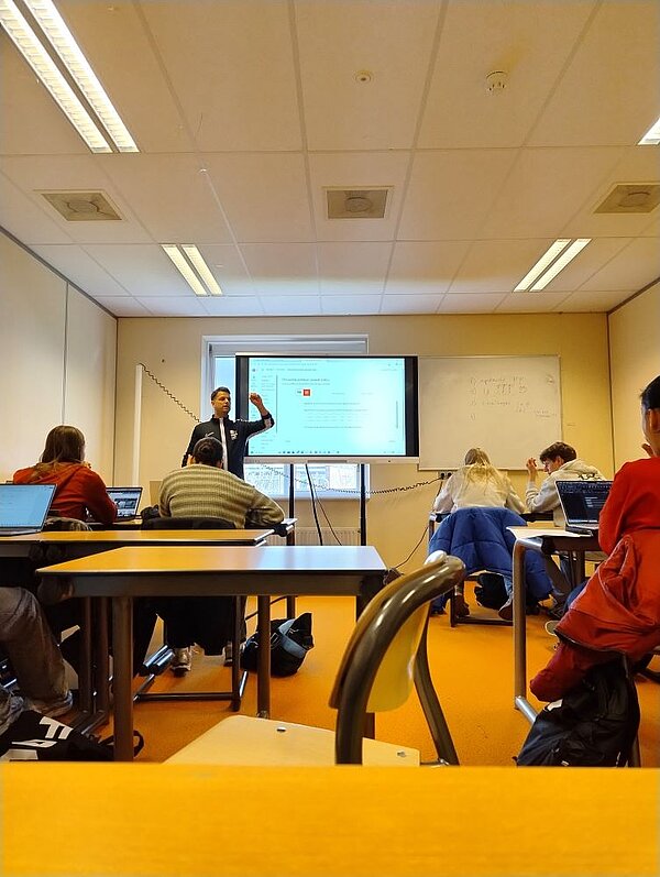 Ein Dozent hält einen interaktiven Vortrag in einem modernen Seminarraum. Die Studierenden sitzen an Tischen mit Laptops und folgen der Präsentation auf einer großen Leinwand.