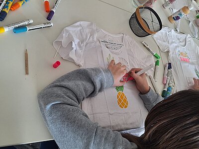 Eine Person bemalt ein weißes T-Shirt mit bunten Stiften und benutzt dafür eine Schablone in Form einer Ananas.