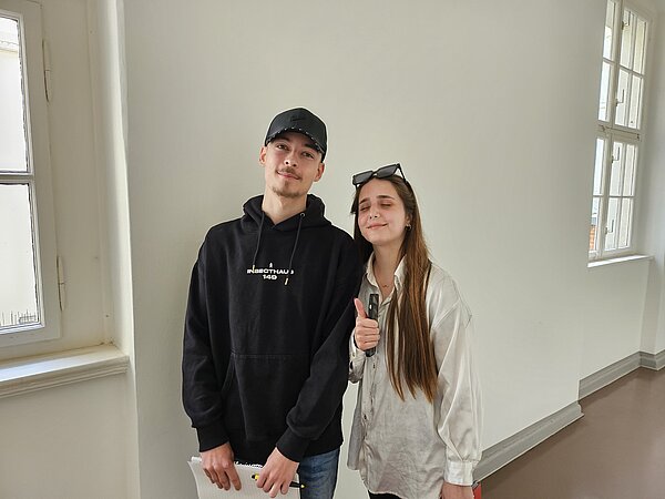 Podcast-Projekt: KAFFEE TRINKEN & SPIELEN Ein Schüler mit schwarzem Hoodie und eine Schülerin mit weiß-grauen Klamotten stehen vor der Kamera und lächeln.era