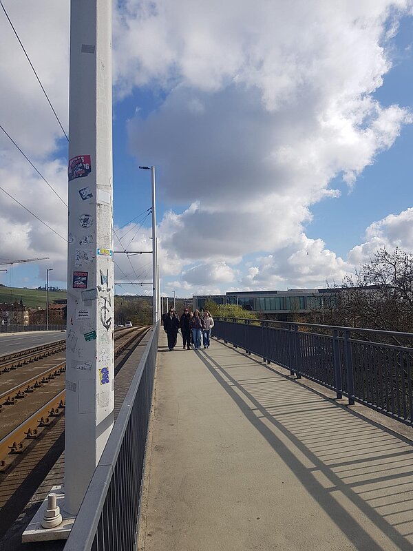 Mehrere Jugendliche gehen bei sonnigem Wetter über einen breiten Fußweg neben Straßenbahnschienen und einer Brücke.