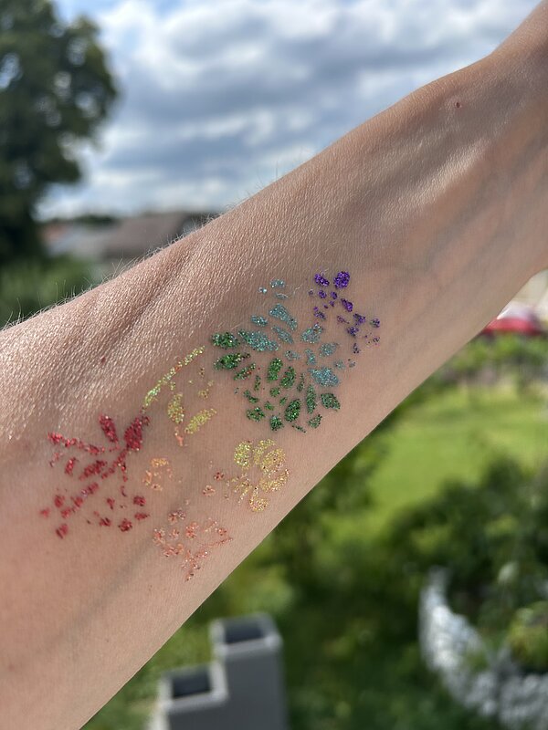 Glitzer-Tattoo auf einem Unterarm, bestehend aus bunten, funkelnden Mustern in Regenbogenfarben. Im Hintergrund Bäume, Wiese und ein leicht bewölkter Himmel.