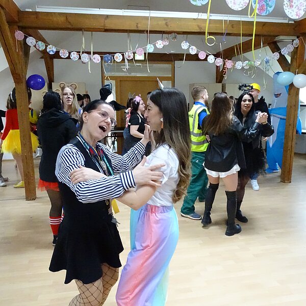 Zwei Frauen tanzen miteinander und lachen. Eine von ihnen trägt einen Regenbogenrock, die andere ein schwarz-weiß gestreiftes Kostüm mit Netzstrumpfhosen. Im Hintergrund tanzen weitere Gäste in bunten Outfits.