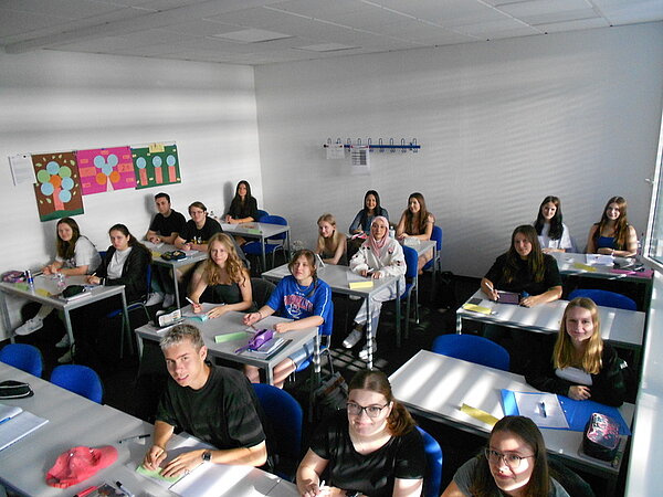 Herzlich willkommen In einem Klassenzimmer sitzen Schülerinnen und Schüler an Schreibtischen. Nicht alle Tische sind komplett belegt. Die Sonne scheint durch die Markisen und wirft Schatten. An der Wand hängen Plakate und eine Garderobe.