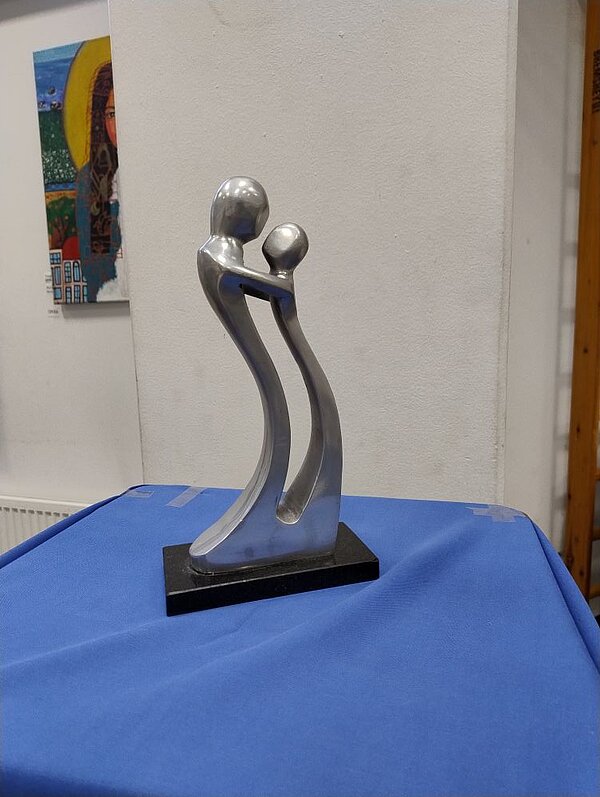 Nahaufnahme einer abstrakten Skulptur aus Metall, die einen größeren und einen kleineren Menschen darstellt, die sich umarmen. Die Skulptur steht auf einer Fläche, die mit blauem Stoff bezogen ist. Im Hintergrund hängt ein buntes Gemälde mit einem Frauenkopf an einer weißen Wand.
