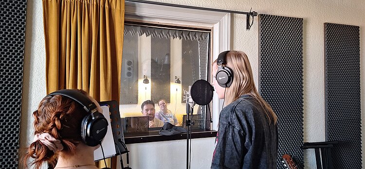 Zwei junge Frauen mit Kopfhörern stehen in einem Tonstudio vor Mikrofon und Notenständer, während zwei weitere Personen sie hinter einer Glasscheibe aus dem Regieraum beobachten.