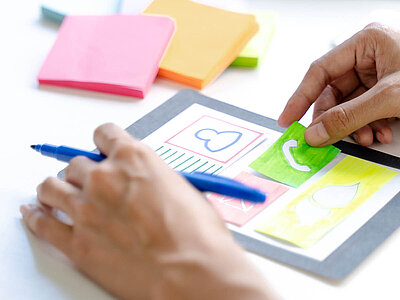 Auf dem Bild sind Hände zu sehen, die bunte Symbole auf Papier zeichnen und dadurch ein Papierhandy kreieren. 