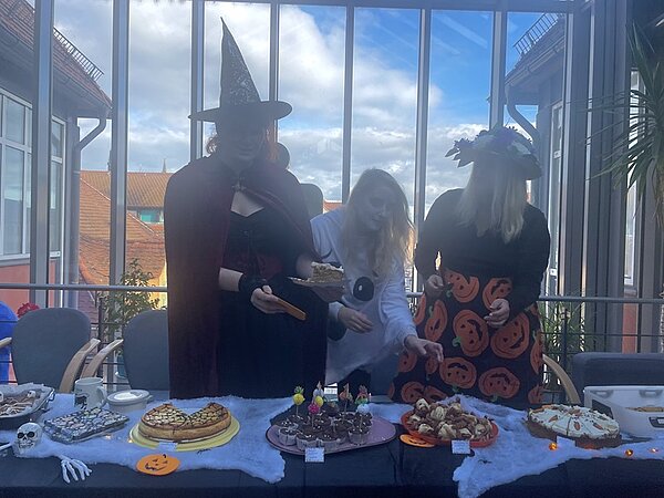 Halloween-Kuchenbasar in der Euro Akademie und den Euro-Schulen in Meißen Teilnehmende am Kuchenbasar stehen hinter der gedeckten Kuchentafel, die in Halloween-Manier dekoriert ist. Die drei weiblichen Teilnehmenden tragen verschiedene Kostüme, unter anderem als Hexe.