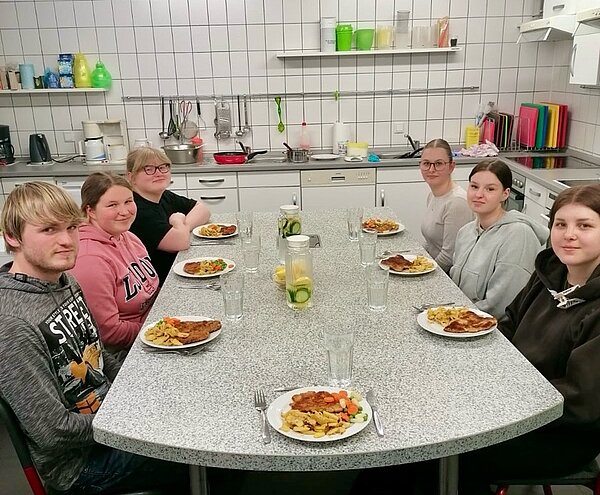 An die Töpfe, fertig, los! Die Auszubildenden sitzen gemeinsam an einem großen Tisch, mit ihrem selbst zubereiteten Gericht.