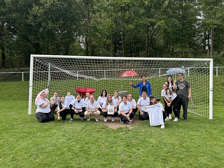 Die Gruppe der angehenden Erzieherinnen und Erzieher steht und hockt vor einem Fußballtor auf einer Rasenfläche und posiert für ein Gruppenfoto. Die meisten tragen weiße T-Shirts, einige halten ein T-Shirt oder ein Blatt Papier in den Händen. Im Hintergrund sind Bäume und ein Zaun zu sehen. Zwei rote Regenschirme werden von Personen gehalten. Die Stimmung wirkt entspannt und gemeinschaftlich.