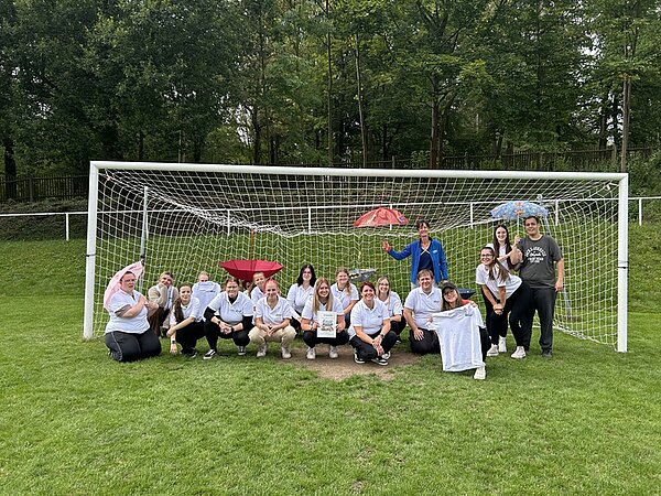 Die Gruppe der angehenden Erzieherinnen und Erzieher steht und hockt vor einem Fußballtor auf einer Rasenfläche und posiert für ein Gruppenfoto. Die meisten tragen weiße T-Shirts, einige halten ein T-Shirt oder ein Blatt Papier in den Händen. Im Hintergrund sind Bäume und ein Zaun zu sehen. Zwei rote Regenschirme werden von Personen gehalten. Die Stimmung wirkt entspannt und gemeinschaftlich.