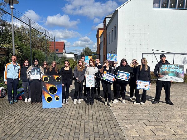 Gruppe von etwa 15 Personen auf einem gepflasterten Platz, mehrere halten selbstgestaltete Plakate mit Weltraum-Motiven wie Raketen und Planeten, darunter ein Schild ‚Operation Mondlandung'.