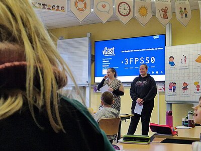 Zwei Personen stehen vorne im Klassenraum und sprechen zu einer sitzenden Klasse; auf dem großen Bildschirm im Hintergrund ist eine Verbindungsseite (vCast) zu sehen.