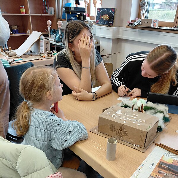 Nahaufnahme einer kleinen Gruppe: Ein Mädchen im Grundschulalter, eine junge Frau mit Hand im Gesicht und eine weitere junge Frau, die sich über ein Kartonmodell beugt und schreibt, sitzen gemeinsam an einem Tisch und basteln.