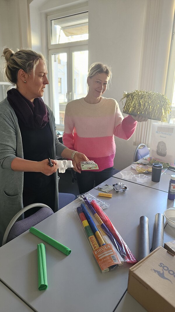Robotik: Innovative Ansätze in der Pflege Zwei Frauen stehen an einem Tisch mit Bastelmaterial. Eine von ihnen hält einen Karton mit goldenem Lametta daran in die Kamera.