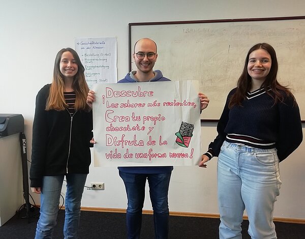 Der nächste Schritt zur Karriere: Career Week Ein Schüler hält ein Plakat mit einer spanischen Werbung in die Kamera, links uns rechts stehen zwei Schülerinnen, die mit zum Team gehören.
