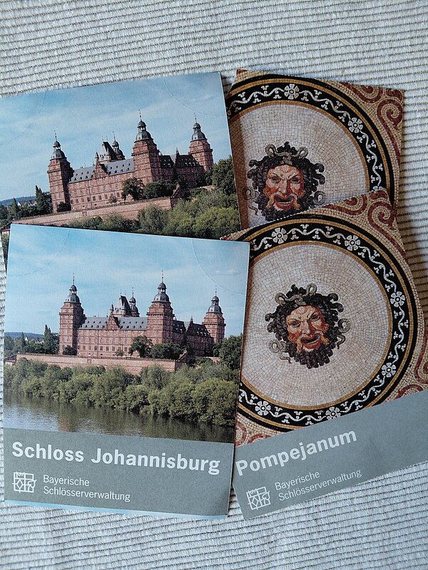 Eintauchen in die Geschichte Aschaffenburgs Zwei Broschüren der Bayerischen Schlösserverwaltung liegen auf einer hellen Oberfläche. Eine zeigt Schloss Johannisburg, die andere ein Mosaik aus dem Pompejanum.
