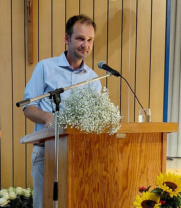 Herz. Humor. Haltung. Stephan Heberlein, Leiter der Euro Akademie Tauberbischofsheim, steht am Rednerpult und spricht lächelnd ins Mikrophon. Auf dem Pult liegen Blumen und steht ein Glas Wasser, auf dem Boden davor steht eine große gläserne Vase mit mehreren Sonnenblumen.