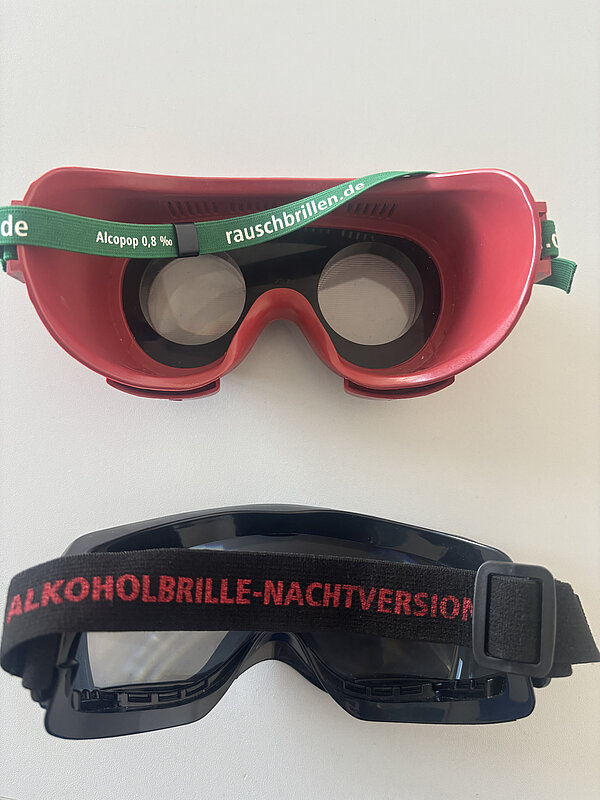 Auf einem weißen Untergrund liegen zwei "Rauschbrillen", die ähnlich aussehen wie Taucherbrillen; die obere ist rot, die untere schwarz.