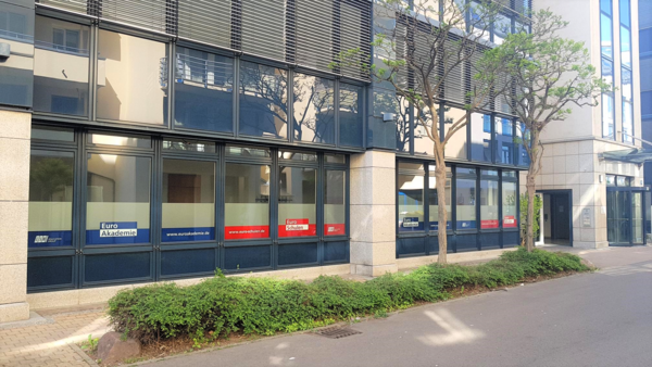 Außenansicht eines modernen Büro- und Schulgebäudes mit großen Glasfronten, Beschriftung „Euro Akademie“ und „Euro Schulen“ auf den Fenstern sowie niedrigen Sträuchern und Bäumen entlang des Gehwegs.